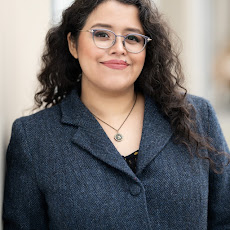 Photograph of the author wearing a formal dark grey blazer, eyeglasses, and a thin necklace. She has curly, dark hair that reaches below her shoulders. She is smiling.