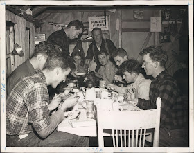 A black and white photograph of a group of men crowded around a dinner table for a meal.