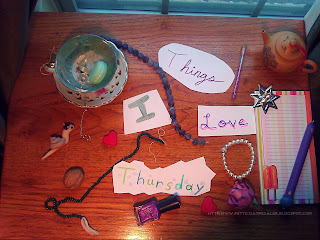 A collection of items arranged as a collage on a small wooden table.  Words written on cut out white paper reads: Things I Love Thursday.  Assorted items in collage include: A mint and white teacup and saucer filled with crystal stones, a quartz pendulum, and a plastic macaroon.  A walnut, two (2) red hearts, a snail wearing a black beret, two (2) strands of stone beads (purple fluorite and green aventurine), a bottle of purple nail polish, a pink rose, a silver colored seven pointed star candle holder, a small tan tea pot, a note pad in summery colors with the image of a popsicle on it, and a decorative pen.  