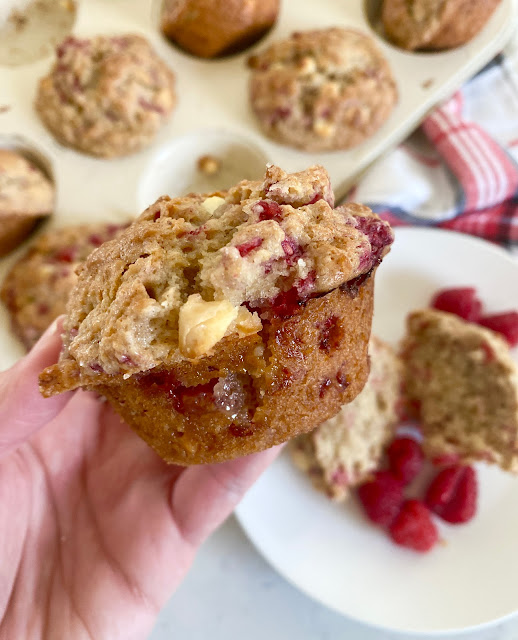 A hand holding a fresh raspberry muffin with a full muffin tin in the background.
