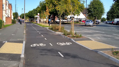 The cycle track with a footway and flats to the left and a planted buffer to the right. There is a pedestrian crossing point with yellowish tactile paving units.