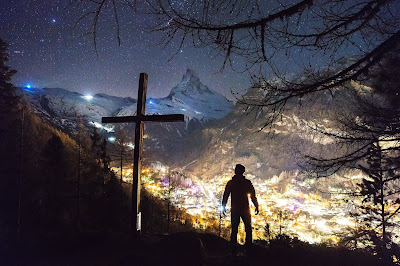 Un giovane in cima a una montagna, accanto a una croce, guarda la città illuminata