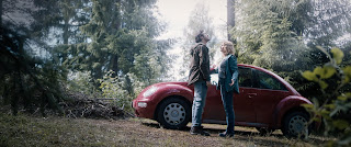 Man and woman in a forest standing in front of a red VW Beetle car