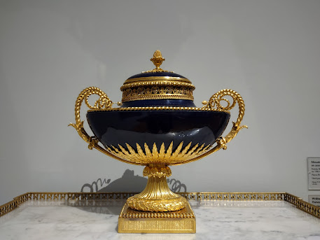 Photo of a black and gold vessel, perhaps a tureen. Square gold pedestal, round fluted gold support, black oval vessel with gold lid and ornate, curlicued gold handles.