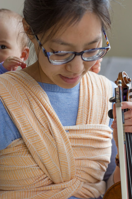 [Image of a smiling tan skin bespectacled Asian woman wearing a toddler on her back in a woven wrap with an undyed combed cotton warp and a mercerized orange weft in a modified herringbone pattern. Mama's looking down at the middle marker tag that happens to be in the center of her freshwater double hammock this time. Mama has violin and bow in hand. Toddler is out of focus and an eye is peeking over Mama's shoulder. Dimpled toddler hands are playing with the wispy dark brown hairs on Mama's neck that don't make it up in the messy bun.]