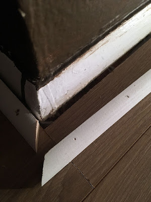 A close-up of the bottom corner of a dark brown wall with two layers of white trim, one clearly part of the wall and one a piece of quarter-round tacked on later. The right-side quarter round has been popped free of the wall, showing the slight gap where the flooring approaches it.