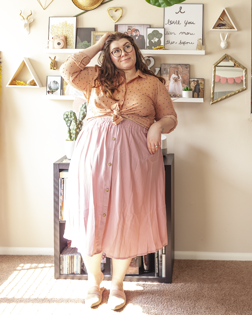 An outfit consisting of a muted pink button down blouse with the sleeves rolled up and tied at the waist and a pink pleated midi skirt and muted pink pointed toe mules.