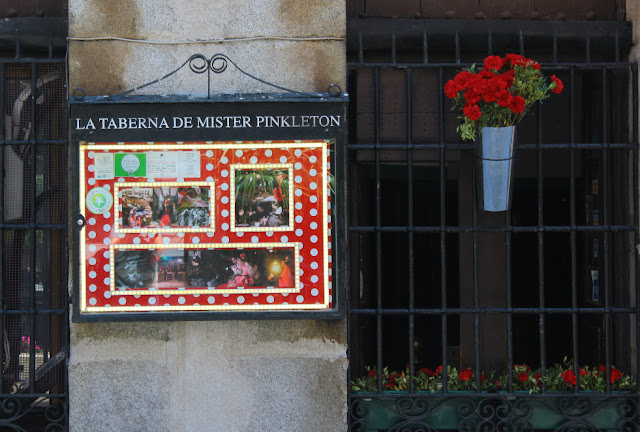 Wejście do baru z muzyką flamenco w Madrycie. Metalowa tablica z napisem La Taberna de Mister Pinkleton. Na tablicy, na czerwonym tle w białe grochy, wiszą zdjęcia z tancerzami flamenco oraz program koncentów. Obok, na zakratowanym oknie, przyczepiona do krat znajduje się aluminiowa donica, w którą włożone są czerwone goździki.