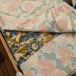 Photo of the inside of the ottoman lid with the top layer of fabric pulled back to reveal another layer of fabric. The top layer is heavy cotton fabric with a pattern of dusky pink flowers on blue-grey background. The layer below is a heavier upholstery fabric of blue and green flowers on a cream background. Both are very old fashioned.