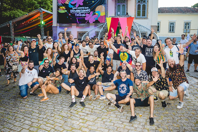 Na imagem, é visto um grupo grande de pessoas vestindo roupas informais, sorrindo e acenando para a câmera diante de um prédio com bandeiras coloridas e um banner onde lê-se Brewing Friends Festival. O ambiente é aberto e ao fundo é visto, do lado esquerdo, uma estrutura metálica comumente usada em eventos com música ao vivo e ao lado direito, uma casa com janelas.  Optou-se por um zoom aberto do flash, sendo possível portanto iluminar todos que posaram para a foto.