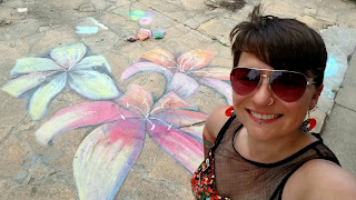 A selfie taken by Haley McAndrews, wearing brown aviator style sunglasses, in front of her sidewalk chalk artwork of three colorful lilies.