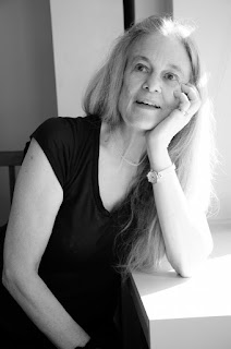 Image of Sharon Olds in black and white. Sharon is sitting by a window with one arm resting on the windowsill. She rests her head on her hand and looks off into the distance, her mouth open as if she is in the middle of a conversation.