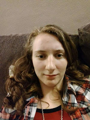 Bonje, a girl with POTS syndrome, lays flat on a brown couch and stares at the camera. Her eyes are tired, her face is pale, and she is barely managing a smile. She is wearing a red and white flannel shirt over a red shirt. Her light brown curls cascade around her neck and shoulders.