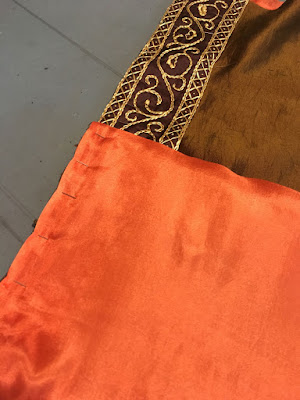 Bright peach silk, pinned at the left edge and folded back to reveal the green-gold silk beneath and the band of gold floral embroidery along the left edge of the green-gold fabric, on a pale blue background.
