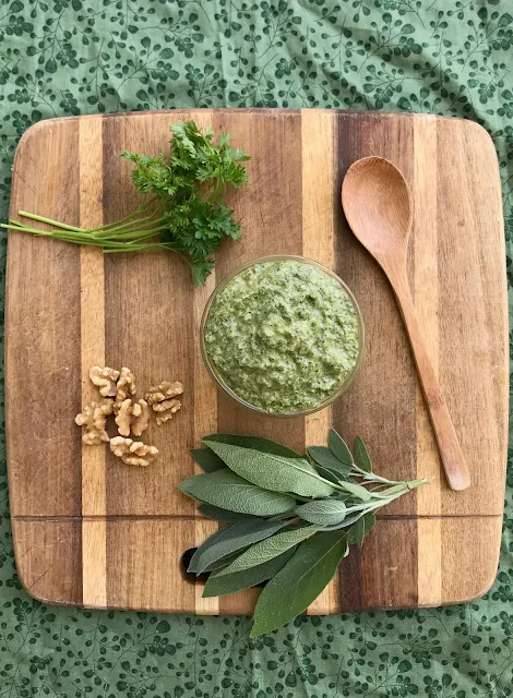 Top view of a jar of parsley and sage walnut pesto on a cutting board with herbs and walnuts.