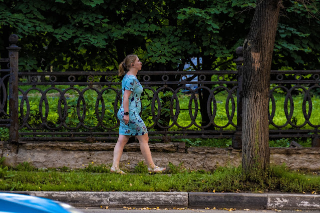 Женщина в голубом платье идет вдоль забора