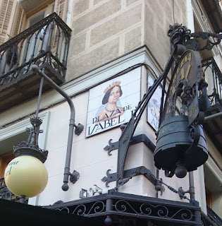 Placa cerámica con el nombre e imagen de la reina, rodeada de adornos de hierro y un farol de un restaurante de la plaza.