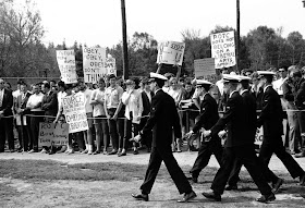 A photograph of a small group of men in uniform walking past a protest.
