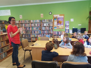 Tło: sala biblioteczna. Pani bibliotekarka stoi na przeciwko dzieci siedzących przy stolikach i pokazuje im książkę.