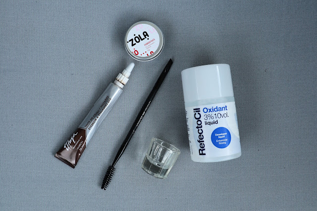 Products used for eyebrow tinging laid out on a grey background. Thuya eyebrow tint, Zola white eyebrow paste, Refectocil 3% oxidant, two-sided brush, and a small container for mixing the products together.