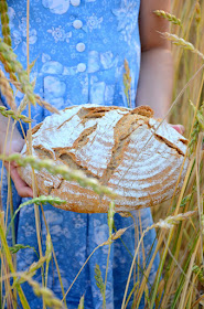 Ein Mädchen in einem blauen Kleid steht in einem Getreidefeld und hält einen Laib Brot in Händen.