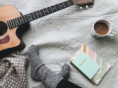 Persona sentada en el suelo con calcetines, una guitarra, cuadernos y una taza de café