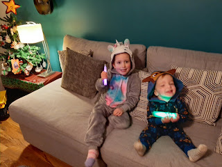 Ellie in a unicorn onesie sits on the couch next to aiden in a paw patrol onesie. Both have glow sticks, it's a party!