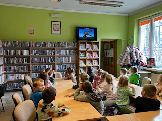 Wnętrze biblioteki. Cała grupa przedszkolaków siedzi przy stolikach. W tle widzimy regały z książkami, regał z czasopismami na którym stoi włączony telewizor. Na telewizorze wyświetlone jest zdjęcie dinozaura.