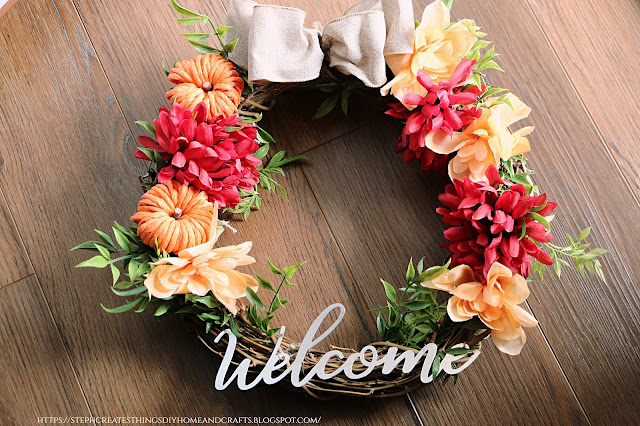 Completed grapevine wreath design with faux yellow and red floral, bow, foliage, and metal welcome sign