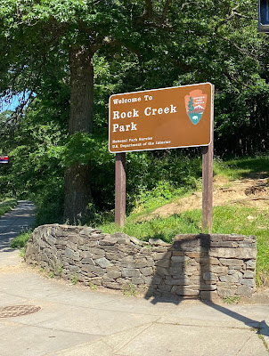 Photo of a stone wall in front of trees, above which is a brown sign reading "Welcome to Rock Creek Park"