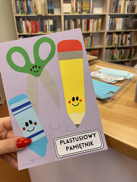 Na pierwszym planie praca plastyczna - na jasnofioletowej kartce przyklejona niebieska kredka, zielone nożyczki, żółty ołówek. Przybory szkolne mają dorysowane czarne oczy i uśmiechy oraz czerwone rumieńce. Za pracą widać ladę biblioteczną. Na ladzie leży pojemnik z papierowymi komponentami, potrzebnymi do wykonania pracy plastycznej. W tle widać regał z książkami.