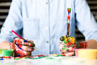 Persona pintando, sosteniendo un pincel y un lápiz, y con las manos manchadas de pintura