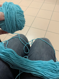 An opened hank of wool lays across the legs of the photographer while they hold a partially wound ball in their left hand. There's a strand of wool from the hank leading to the ball.