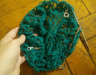 Knit lace on a circular needle.  The yarn lace yarn in a deep blue-green colour.  There are 3 feather stitch markers and 1 dolphin stitch marker clipped into the work.