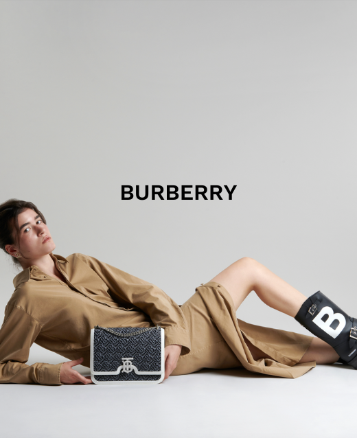 Image of female modeling lying on the ground on her side, she wears a tan coloured shirt dress, with burberry chunky boots and burberry bag in front. She is situated infront of a platinum coloured backdrop and text above is in black and states 'BURBERRY'