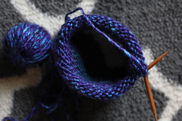 View from above at jewel-toned blue and turquoise knit project on a set of circular bamboo knitting needles against a gray and white rug.