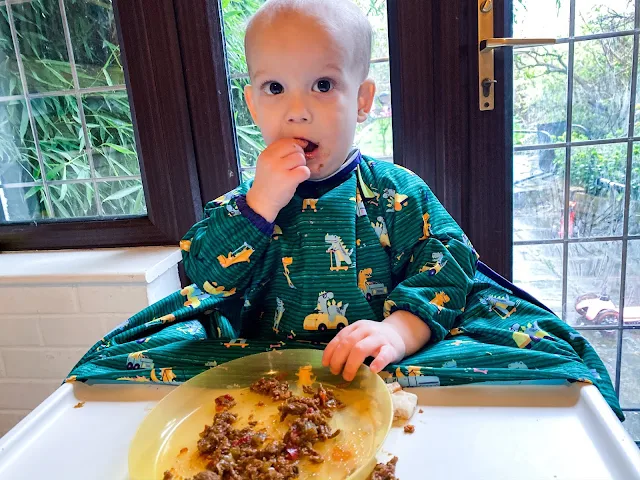 Baby eating food in a green dinosaur coverall all BIBaDO bib