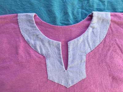 Close up of the neck of a tunic with a decorative facing that has been whip stitched to the right side of the garment. The body of the tunic is bright pink and the facing is lavender. The facing is about 1.5 inches wide around the collar and neck slit, and tapers to a shallow point about 1.5 inches from the bottom of the neck slit. End ID.