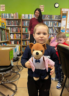 Sala biblioteczna. Chłopczyk z czarnej bluzie i czarnych spodniach pozuje do zdjęcia trzymając w ręku maskotkę misia uszatka. W tle stoją kartonowe pudełka na segregację odpadów, za chłopcem stoi pani bibliotekarka na tle regałów z książkami.