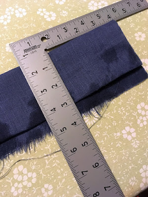 A blue strip of linen, folded in half lengthwise and with the upper edge folded under to make the upper portion three inches wide, according to the metal ell square arranged on top of the strip, all on a pale green floral ironing board.