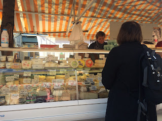 A cheese vendor in the Cours Saleya, with a striped awning overhead, and Mary (from the back) ordering cheese at the counter.