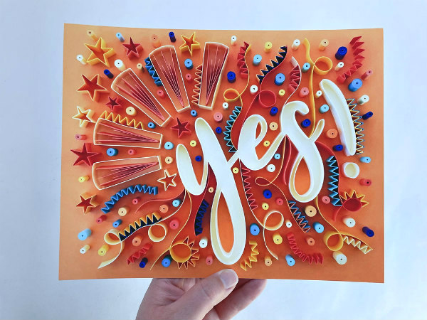 hand holding colorful quilled word yes surrounded by festive on-edge paper stars, squiggles, and dots