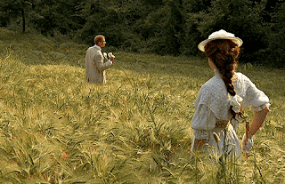 A scene from the 1985 film, A Room With a View. Lucy Honeychurch (Helena Bonham Carter) stands in a field. Ahead of her, turning to see her, is George Emerson (Julian Sands). They are dressed in Edwardian clothes.