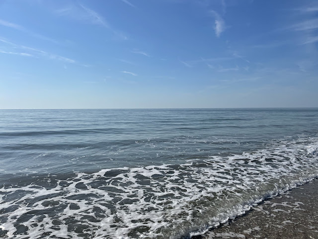 The open ocean with not a human or boat in sight. The ocean extends to the horizon with its dark blue waters. There are white bubbles in the forefront of the photo where the waves are meeting the beach. the water is so calm the ocean almost looks like a lake.