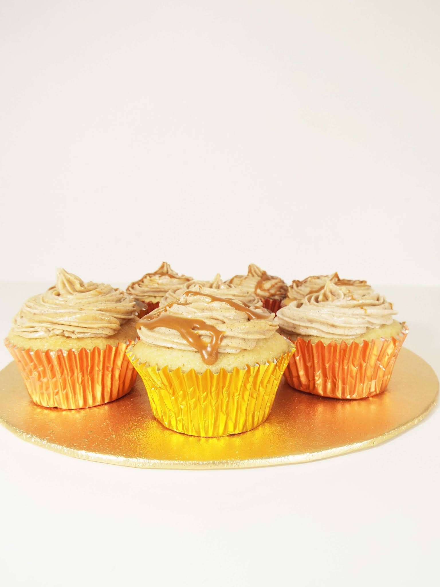 Portrait picture of spread of caramel coloured, iced cupcakes, dressed in copper and gold cases and sat on a gold cake stand. Biscoff sauce if drizzled over the first centre cupcake.