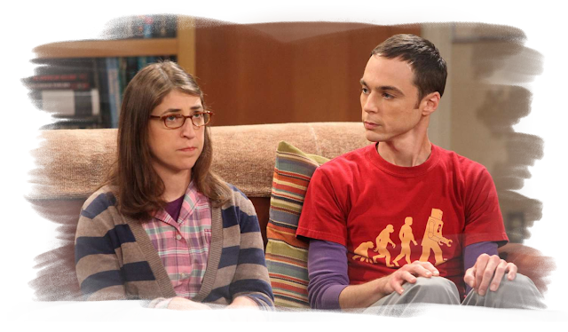 A screenshot from Big Bang Theory of Amy and Sheldon sitting on a couch, where Amy looks annoyed towards someone off camera, while sheldon looks at her, confused.
