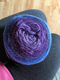 A yarn cake photographed from the top showing rings of several purple shades, gray, and two shades of blue. The entire yarn has a strand of metallic silver run through it.