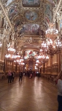 Photograph of a grand public space at the Opera Garnier, Paris. A triple-height room,  with parquet wooden floors, enormous chandeliers, many painted panels on the ceiling.