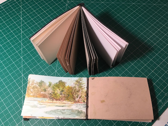 New homemade sketchbook with pages fanned out, next to old sketchbook open to watercolor of trees and icy pond.