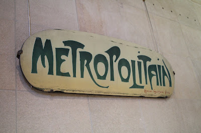 Rectangular cream-colored metal sign with rounded edges saying Metropolitan (as in the Paris subway) in a stylized green font.